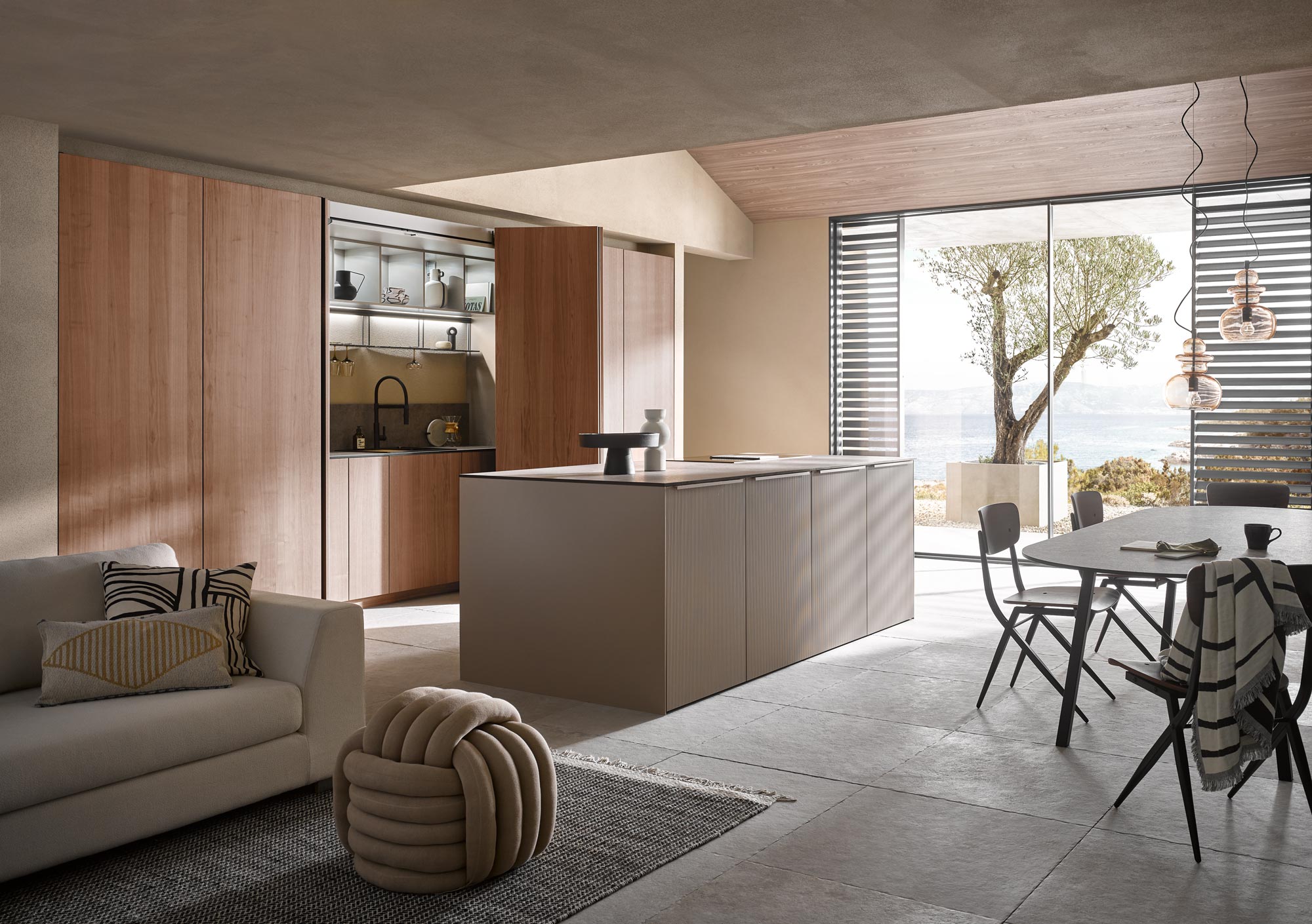 Moderne Küche und Essbereich mit Holzakzenten, Kochinsel, offenen Regalen, neutraler Einrichtung, einem hellen Sofa, einem runden Sitzkissen und großen Fenstern mit Blick auf einen hellen Außenbereich mit Baum und Wasser – ein Beispiel für durchdachte Küchenplanung, die Funktionalität und zeitgenössischen Stil perfekt miteinander verbindet.