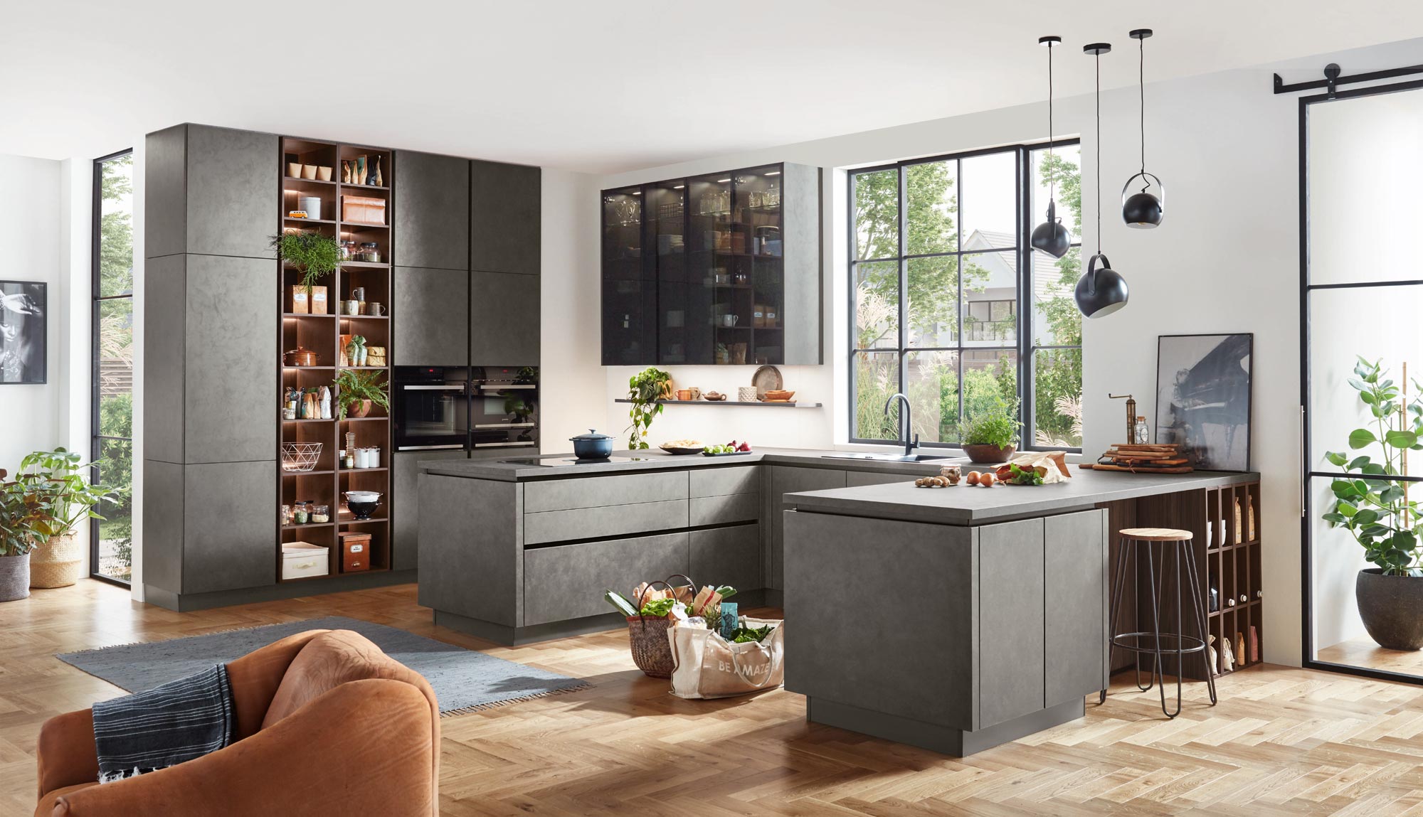 Eine moderne Küche mit grauen Schränken, Holzakzenten und großen Fenstern, durch die viel Tageslicht hereinströmt – perfekt für jede Küchenplanung. Topfpflanzen, eine Kücheninsel mit Hockern, Hängeleuchten und offene Regale schaffen einen gemütlichen, stilvollen und einladenden Raum.